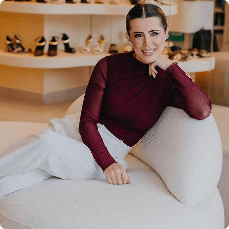 Mulher sentada em um sofá azul em frente a uma janela panorâmica, usando roupa social e expressando postura confiante.